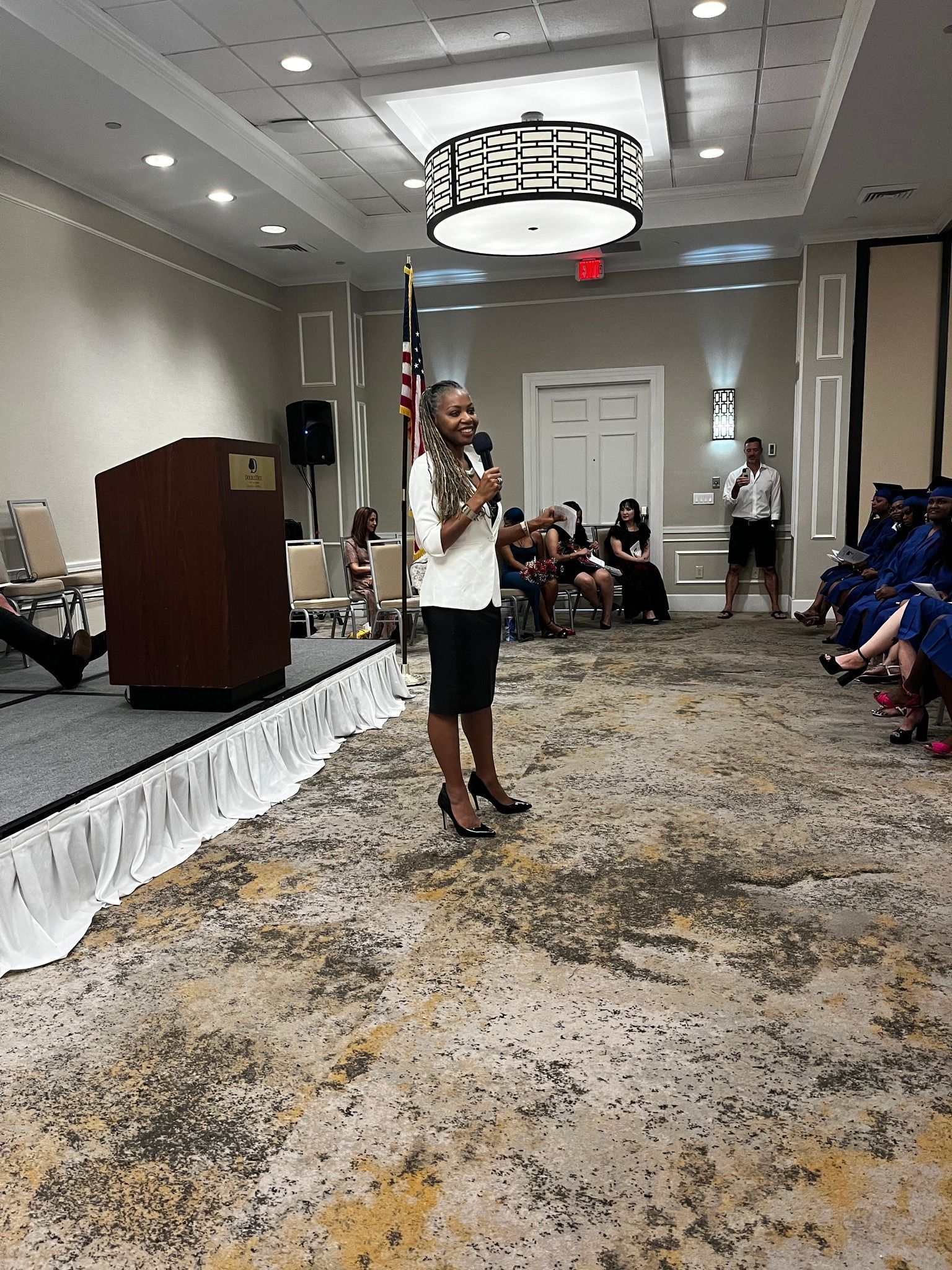 Woman speaking at podium, in formal setting, audience seated, some in graduation gowns.