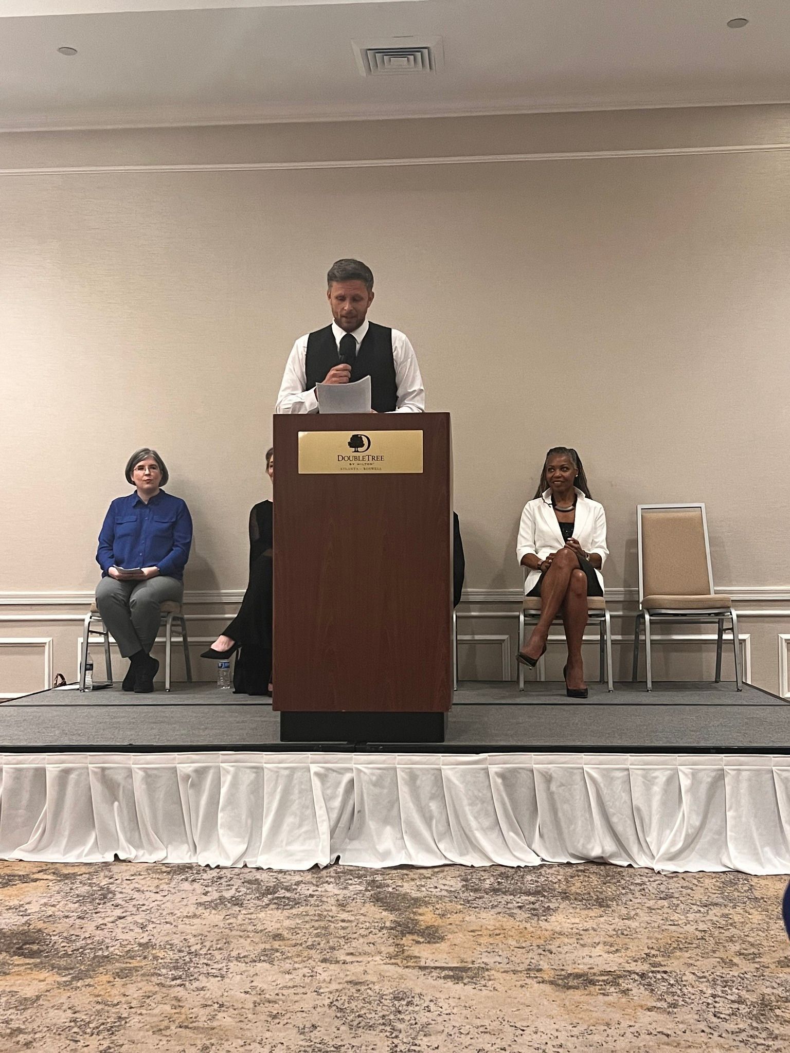 Man at podium speaking; two people seated on stage. Neutral background, hotel setting.
