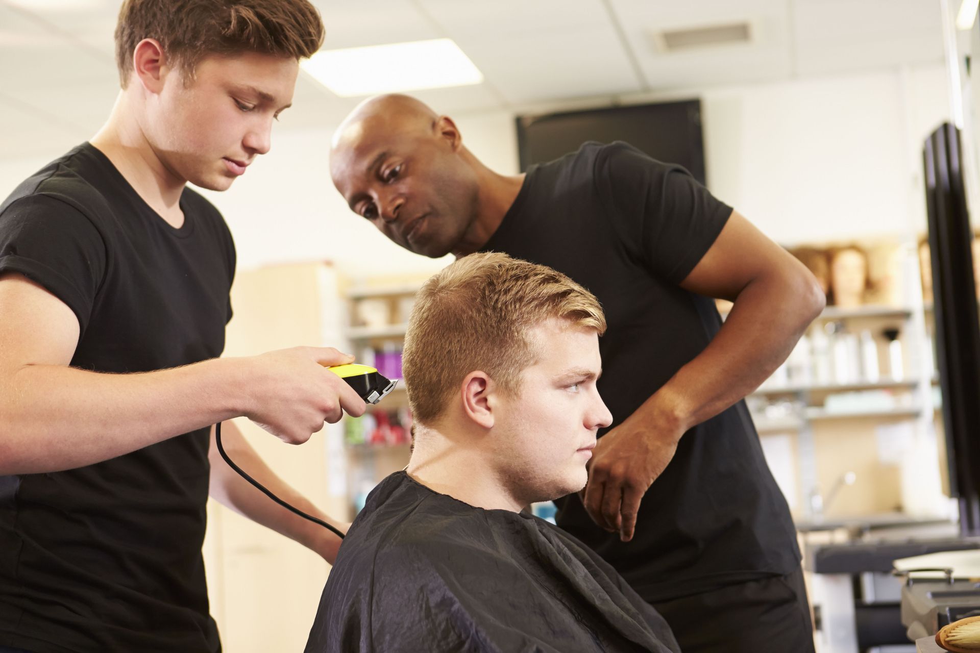Two barbers giving a client a haircut in a salon, one using clippers, the other looking on.