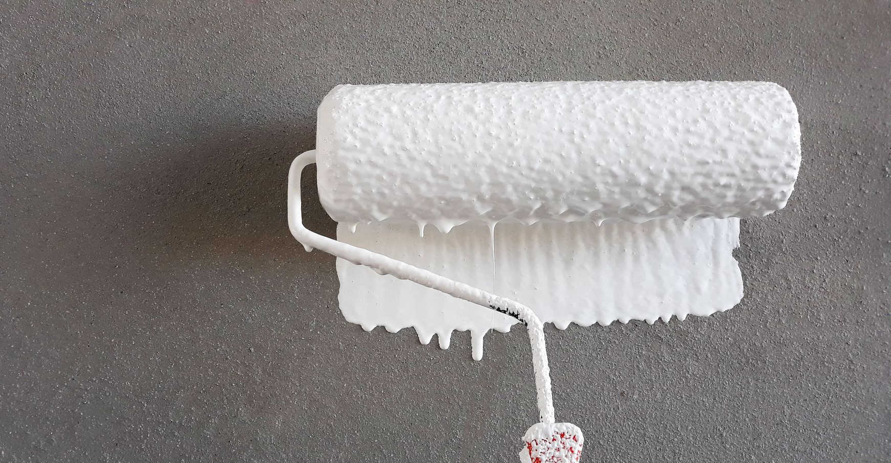 A white paint roller applying white paint to a textured gray wall.