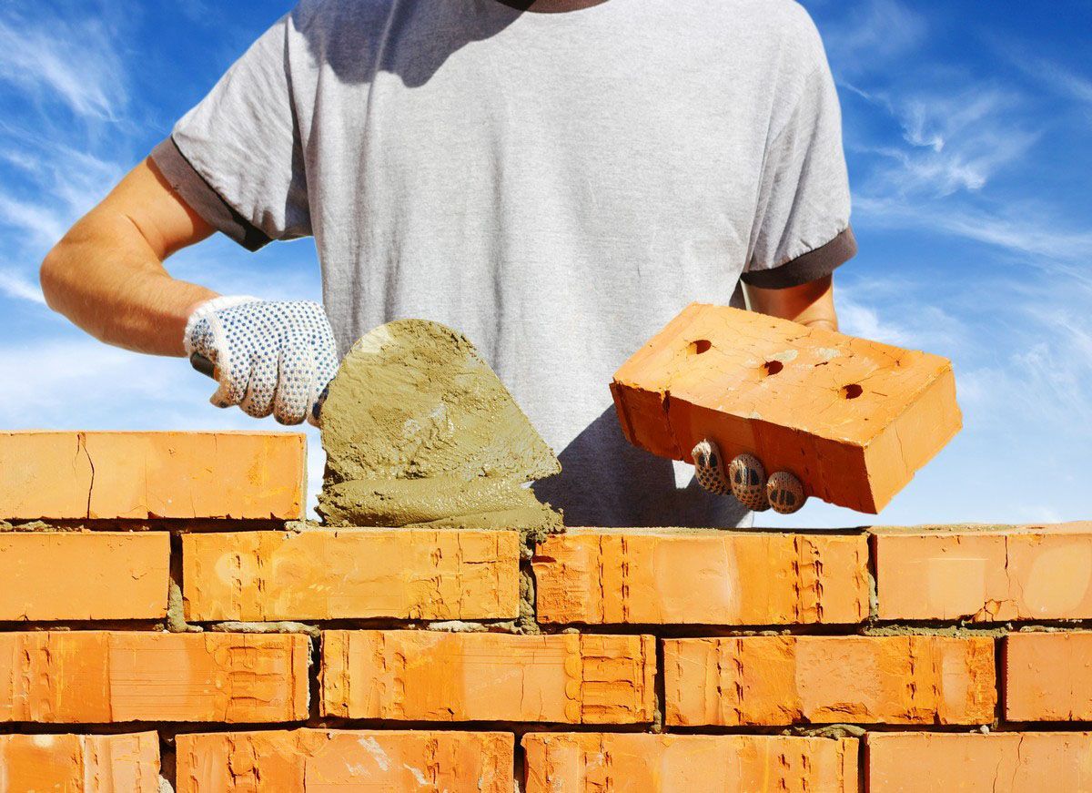 Person laying brick wall, using trowel to apply mortar, blue sky background.