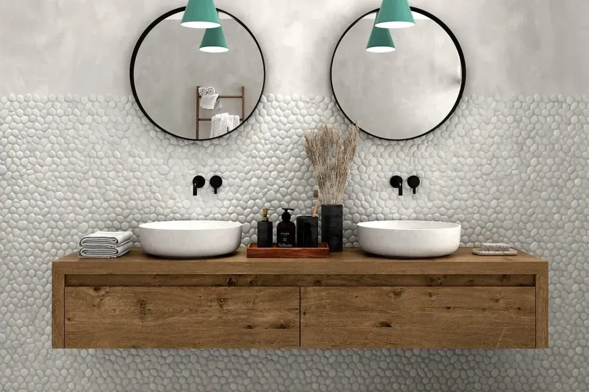Modern bathroom with double sinks, pebble tile wall, wooden vanity, round mirrors, and teal pendant lights.