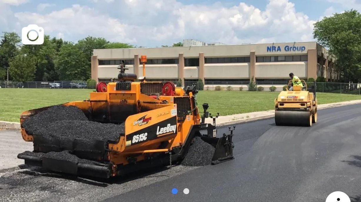 Asphalt paving with a machine laying down black pavement, a worker operating a steam roller, building in background.