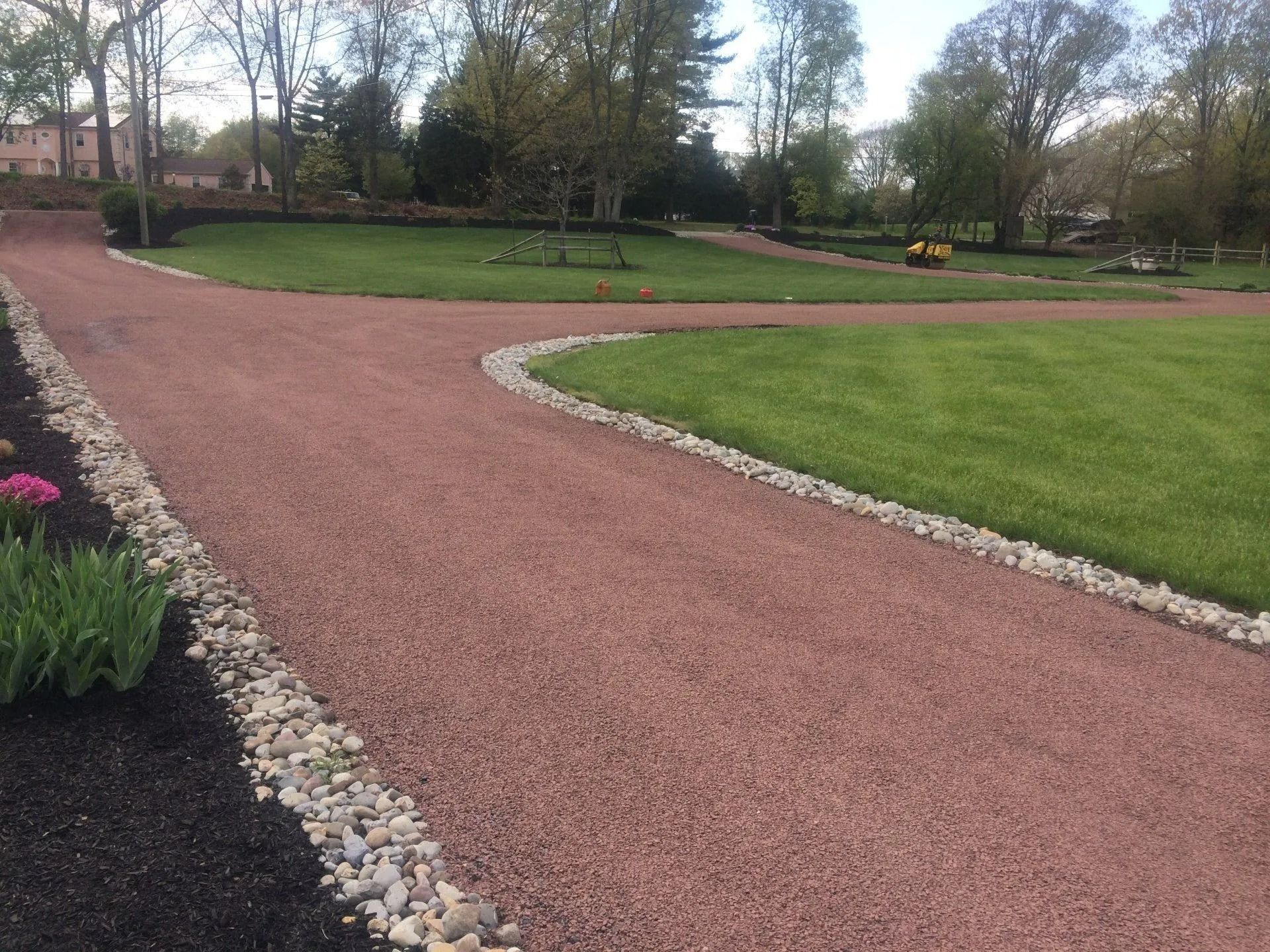 Red gravel driveway curves through green lawn, bordered by white stones and dark mulch.