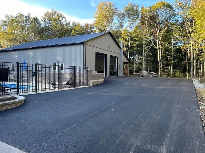 A large, new asphalt driveway leads to a tan metal building with garage doors. Trees and a pool are nearby.