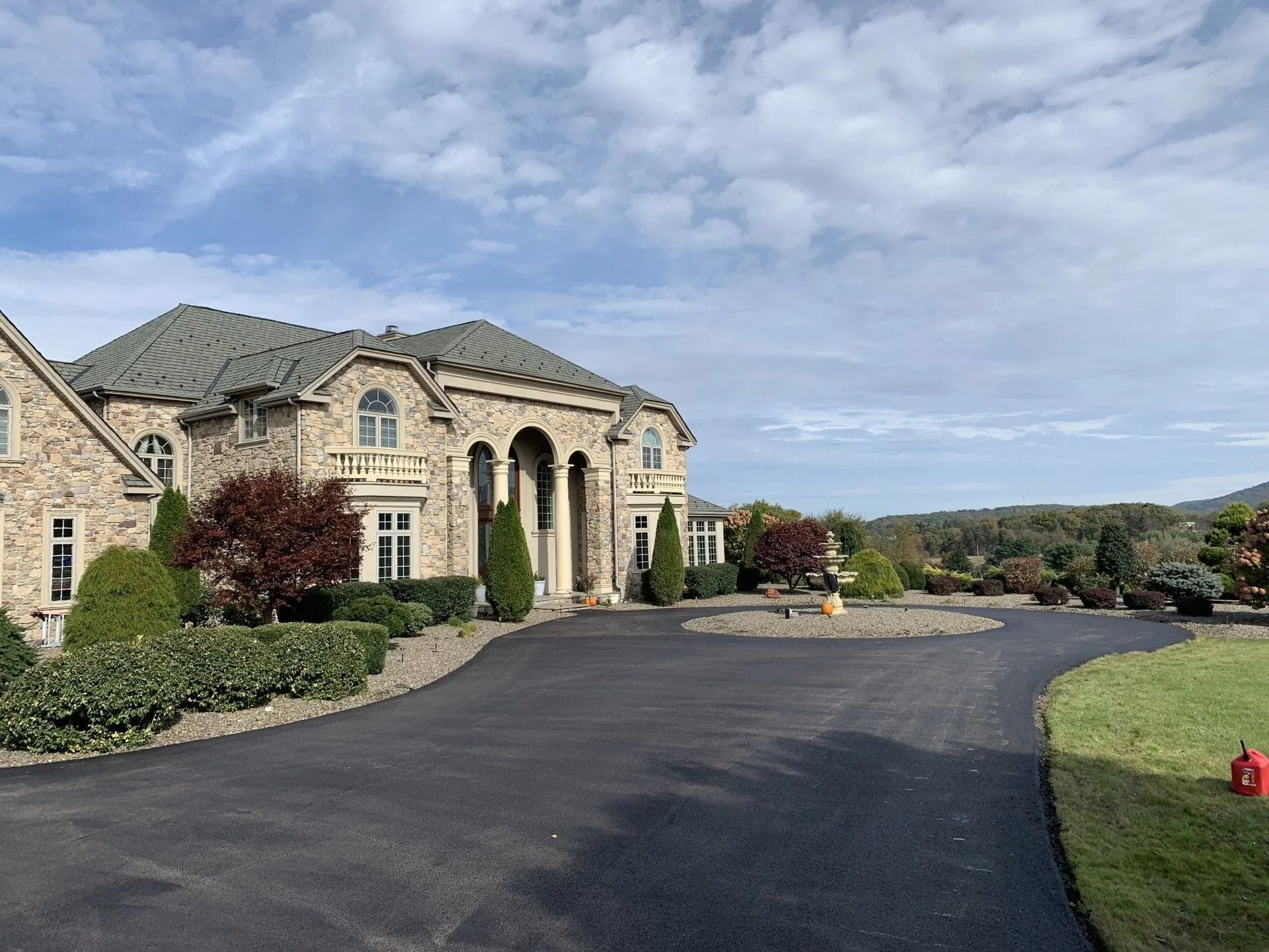 Large stone house with arched entrance, long asphalt driveway, and landscaped yard under a cloudy sky.