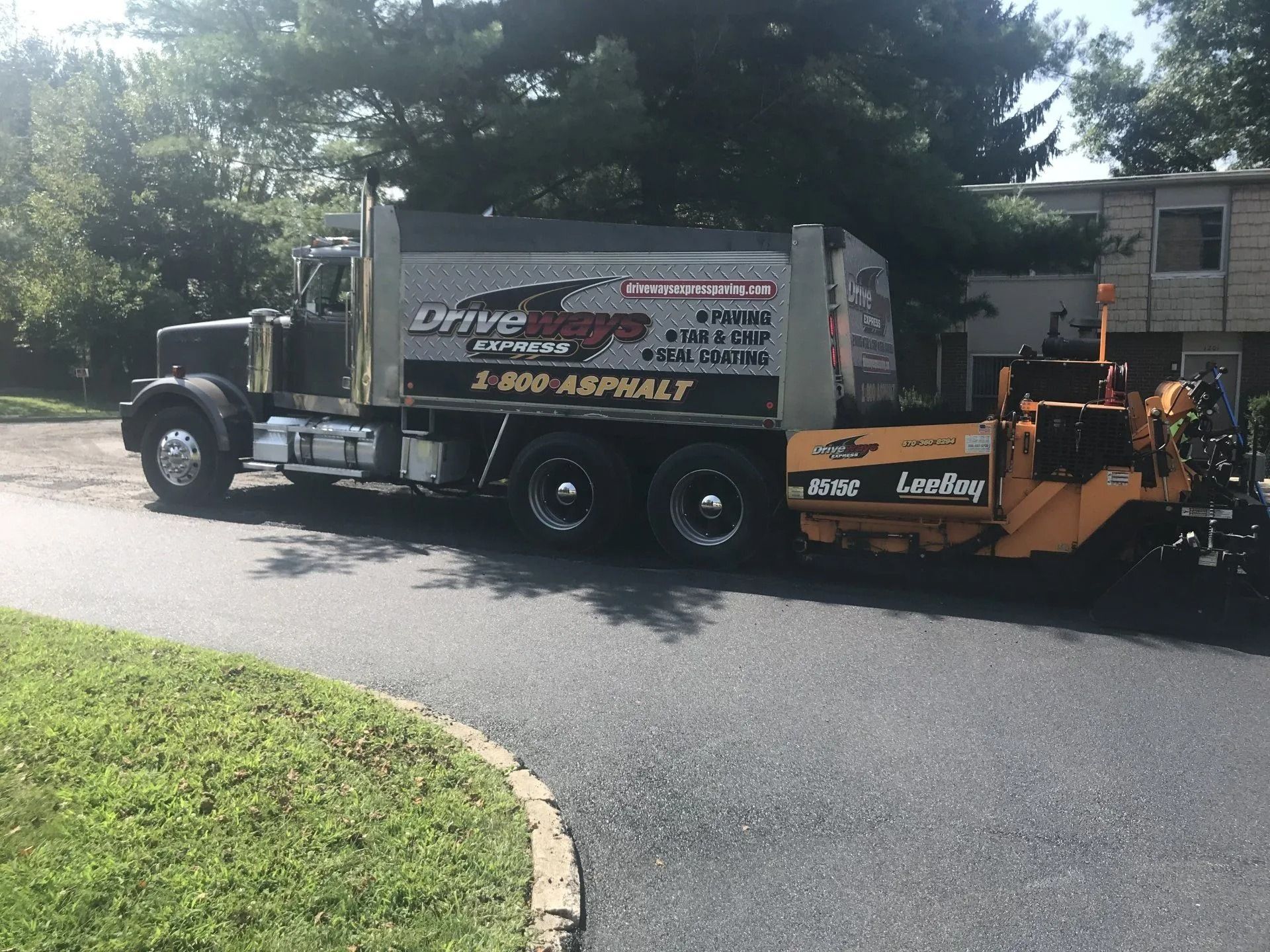 Asphalt paving truck and machinery on a driveway, near a building with a dark asphalt surface.