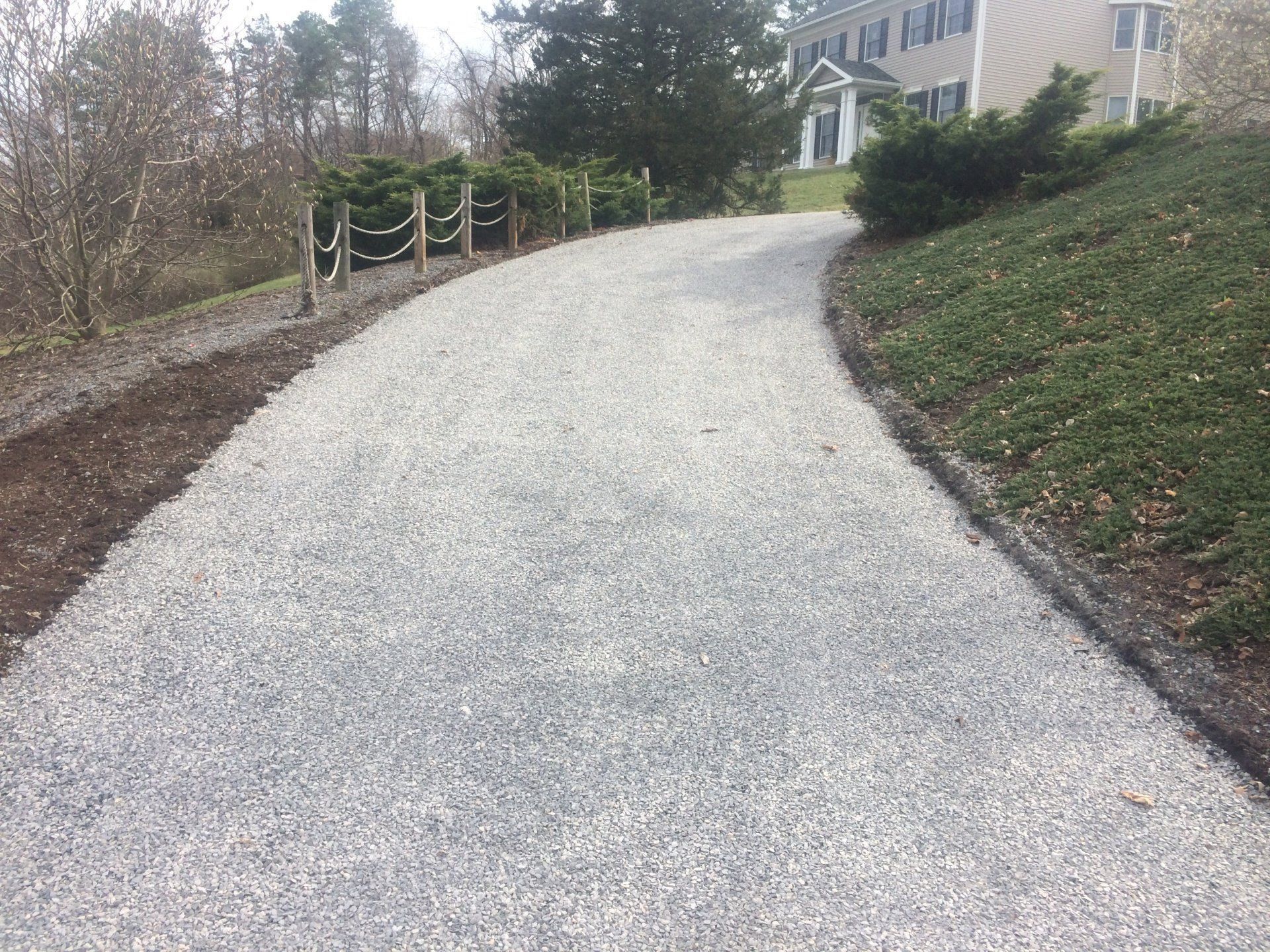 Gravel driveway leading to a house on a slight incline, bordered by landscaping and a wooden fence.