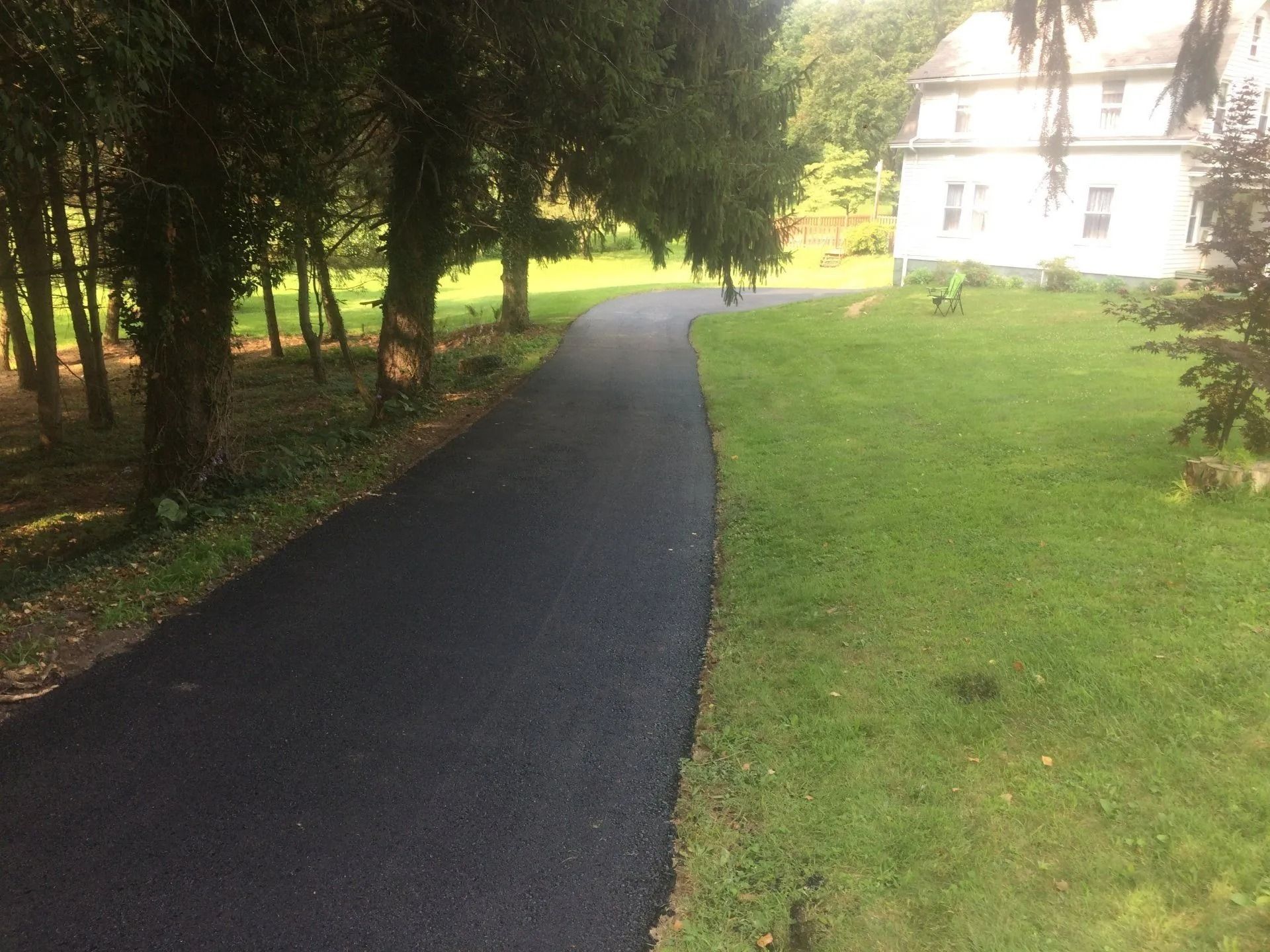 Asphalt driveway curves towards a white house, bordered by grass and trees on a sunny day.