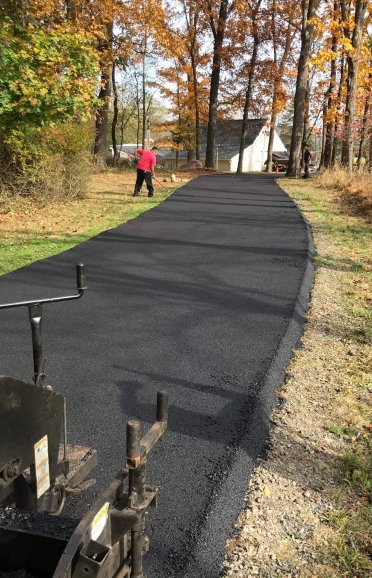 Freshly paved asphalt driveway; a worker in red stands in the distance. Autumn trees frame the scene.