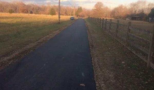 Paved path through a grassy field, bordered by a fence and trees under a hazy sky.