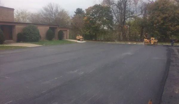 Asphalt paving, two yellow rollers compact new black surface.  Building and trees in background.