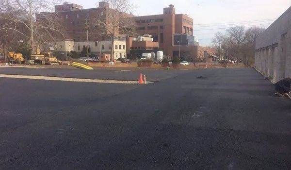 Newly paved asphalt lot with orange cone; buildings in the background.