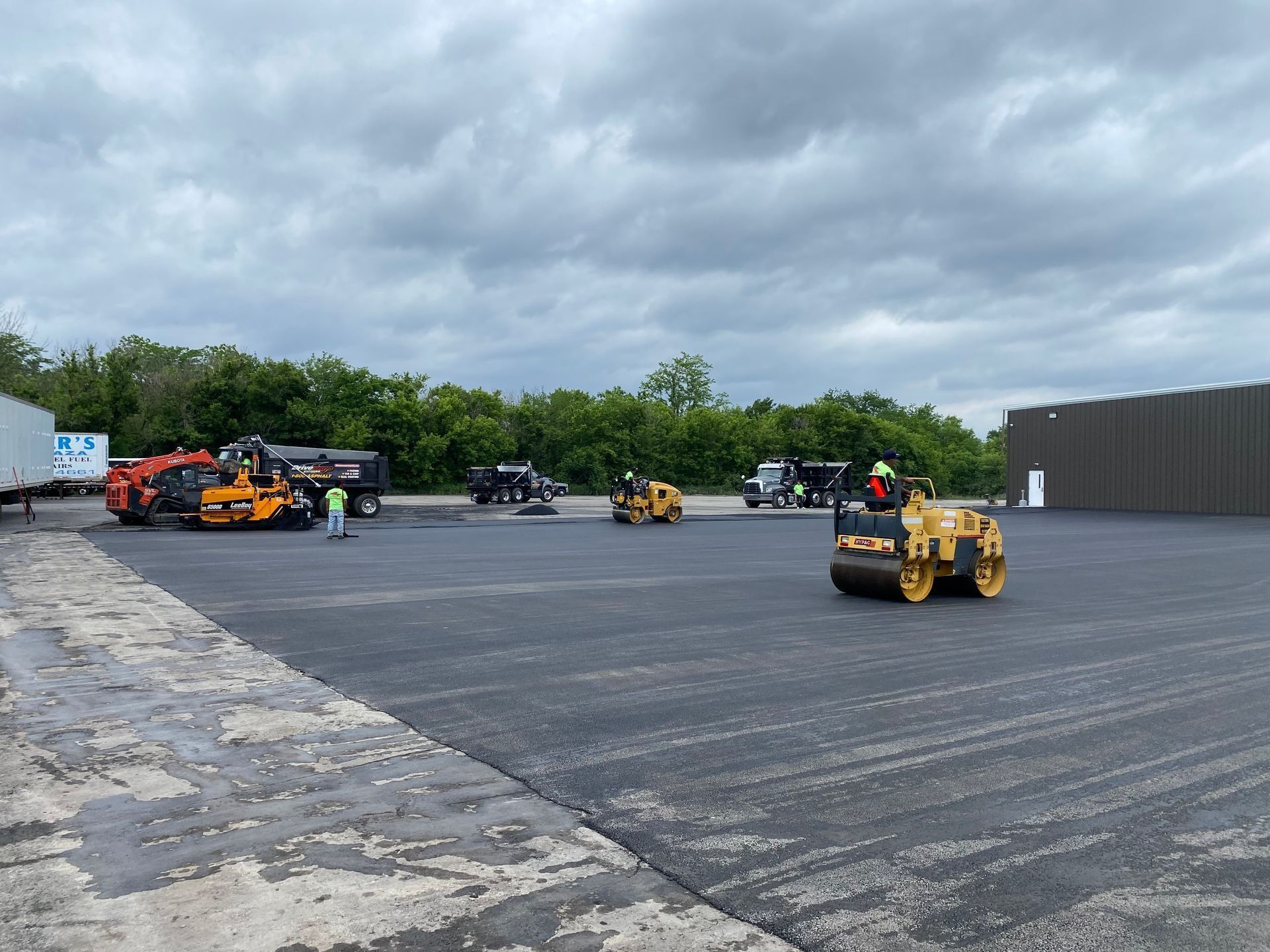 Asphalt paving in progress: rollers, trucks, and workers on a freshly paved area under a cloudy sky.