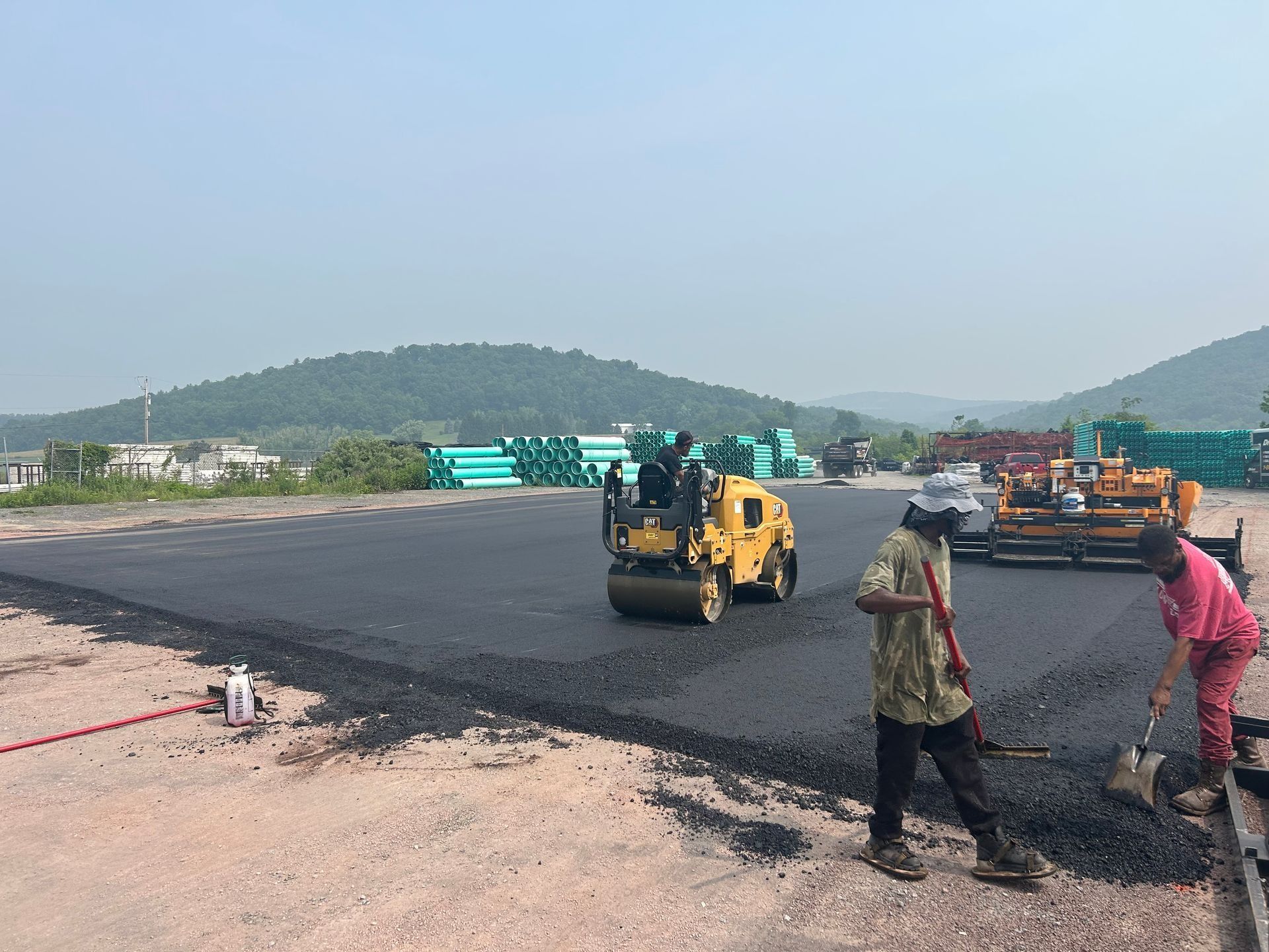 Workers paving a road with a yellow roller and shovels; mountains in background.
