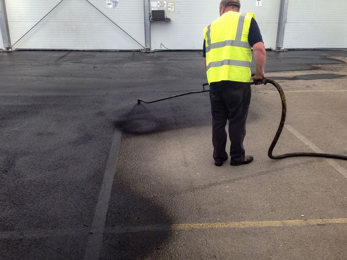 Person in safety vest spraying dark sealant onto asphalt surface.