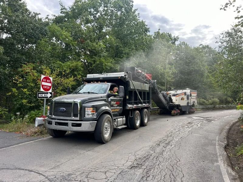 Black truck and road milling machine on a road, preparing for paving. Trees in background,