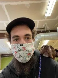 Person in a cap and patterned mask with a brown dog peeking over their shoulder in an indoor space