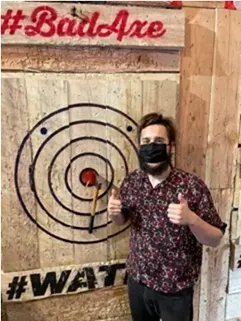 Person giving thumbs up beside an axe-throwing target in a wooden room with #BadAxe and #WAT signs