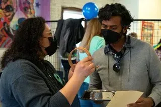 Two masked people talking at an indoor booth, one holding a ring-shaped item and papers.