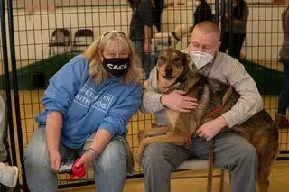 Two people sitting with a large dog in a kennel, one holding a red leash and both wearing face masks.