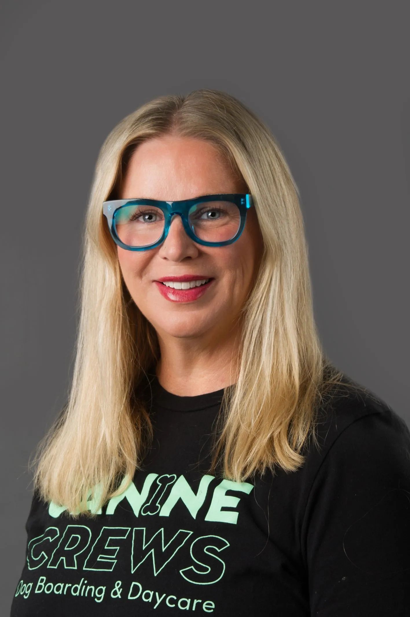 Smiling person with long blonde hair and blue glasses wearing a black T-shirt against a gray background