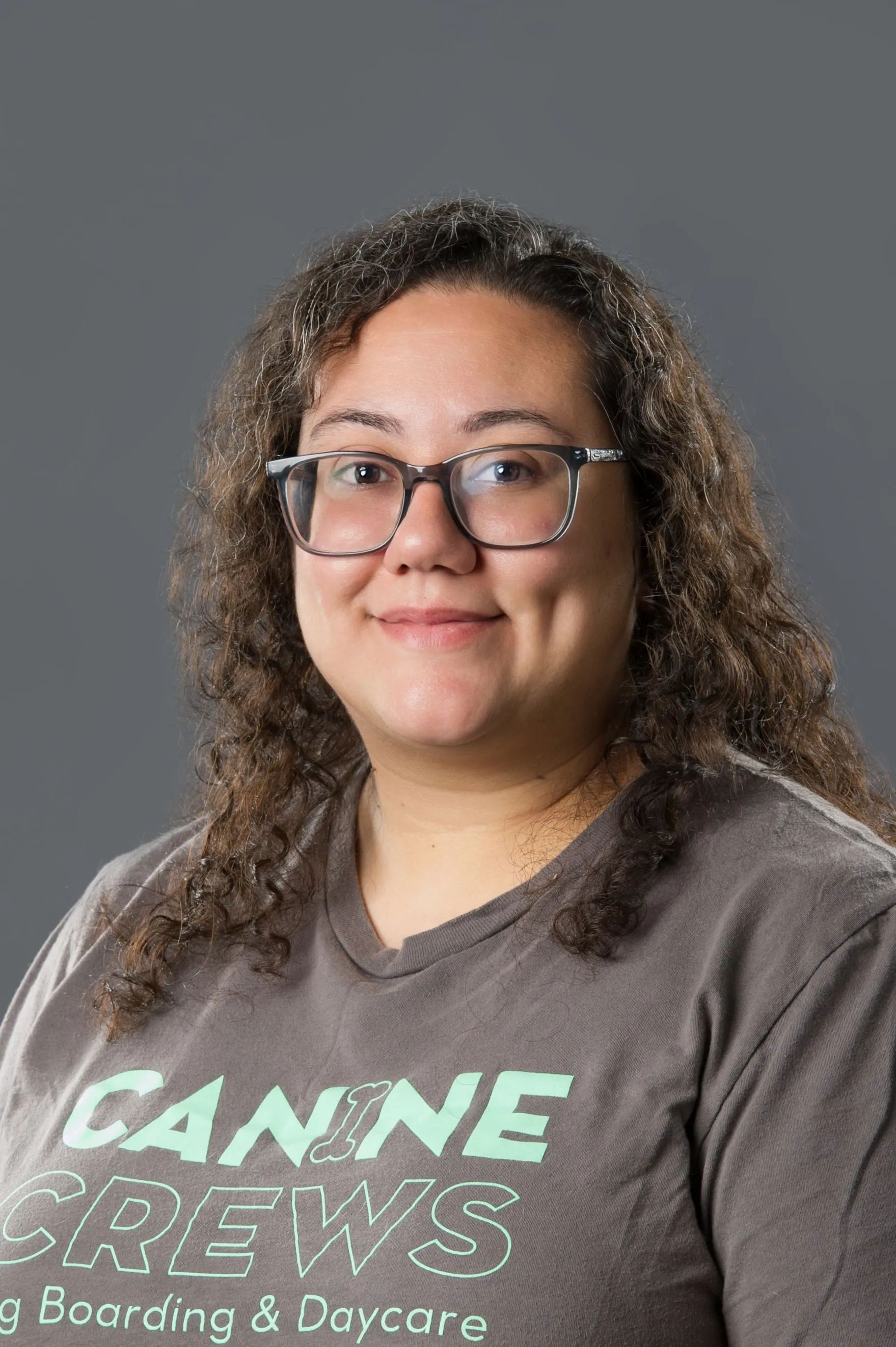 Person with curly hair and glasses wearing a gray “CANNIE CREWS” T-shirt against a gray background