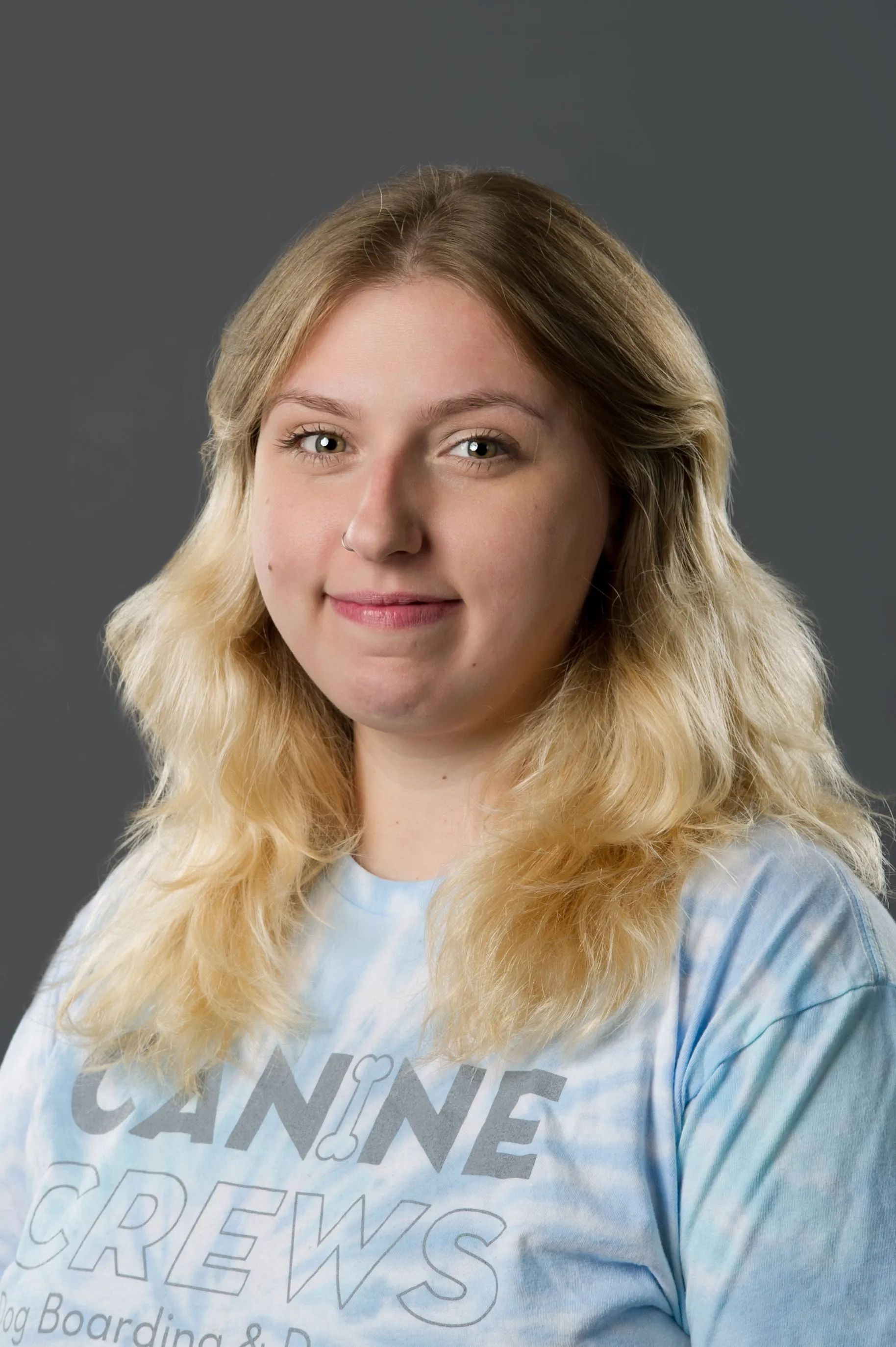 Person with wavy blonde hair in a tie-dye shirt, standing against a gray background.