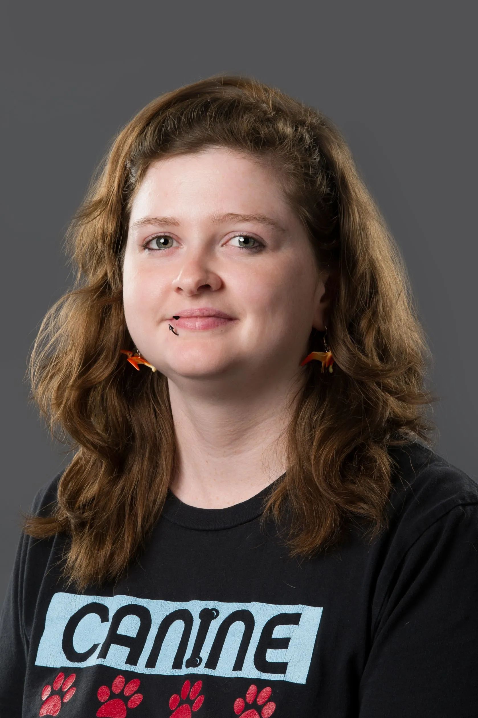 Person with curly brown hair wearing a black “Canada” T-shirt and orange earrings against a gray background