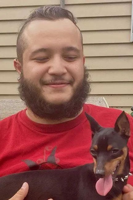Man in red shirt holding a small black-and-tan dog with its tongue out, standing outdoors by siding