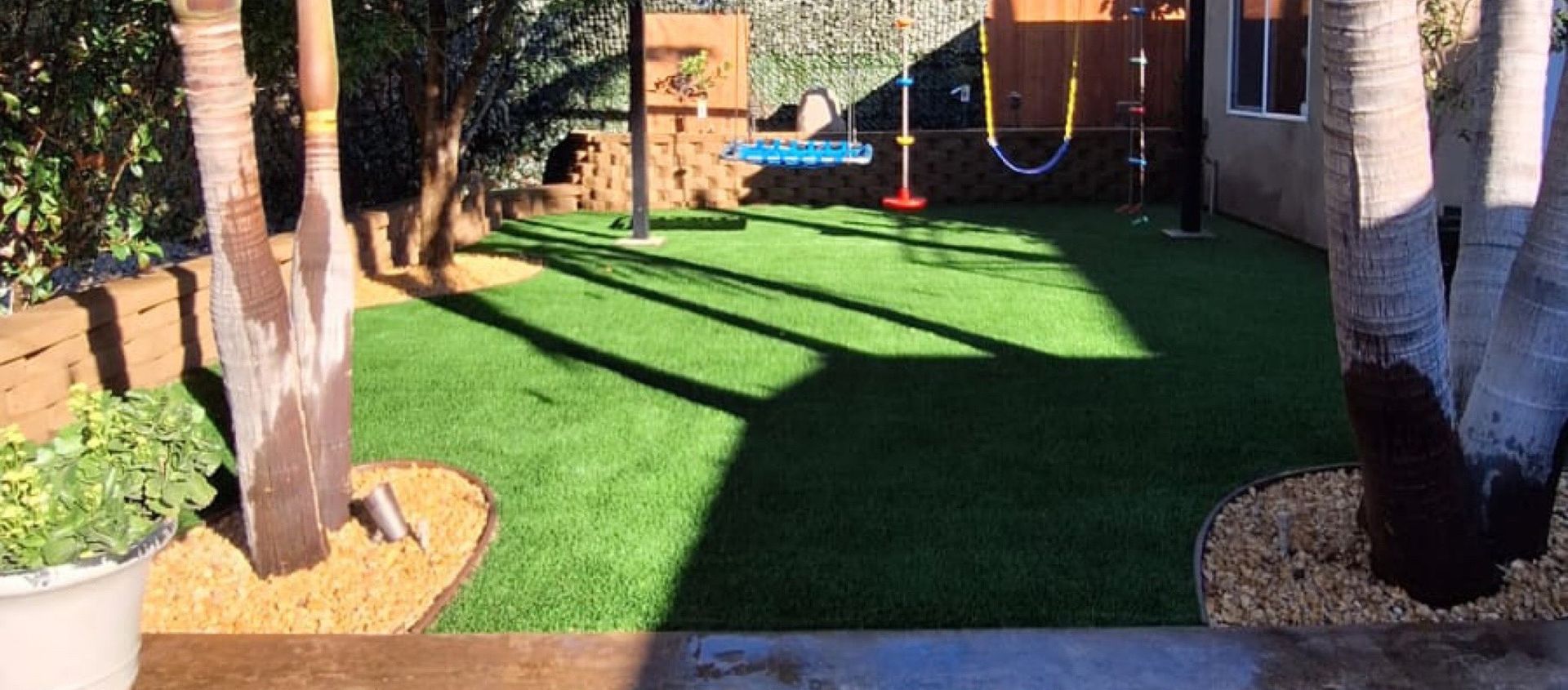 A backyard with dead grass, trees, a stone wall, and a wooden fence - after.