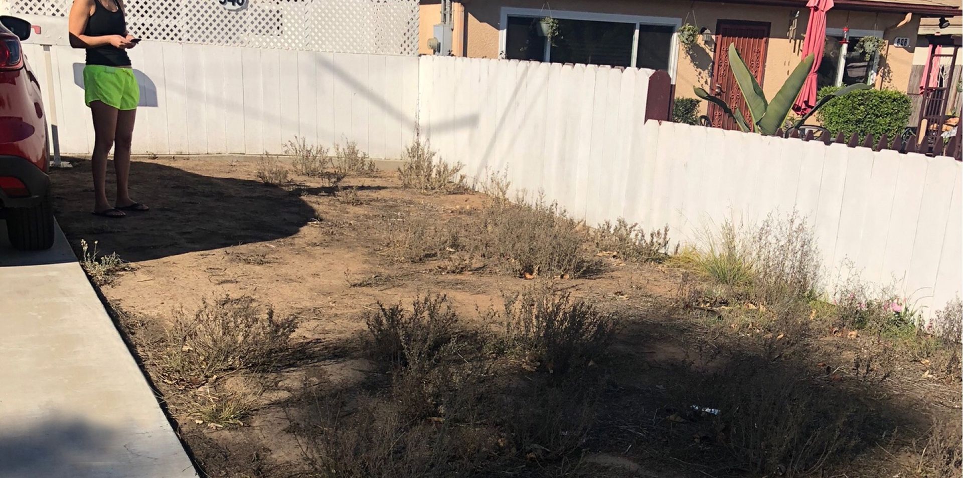 Person in green shorts standing near a car and a dirt patch in front of a white fence - before.