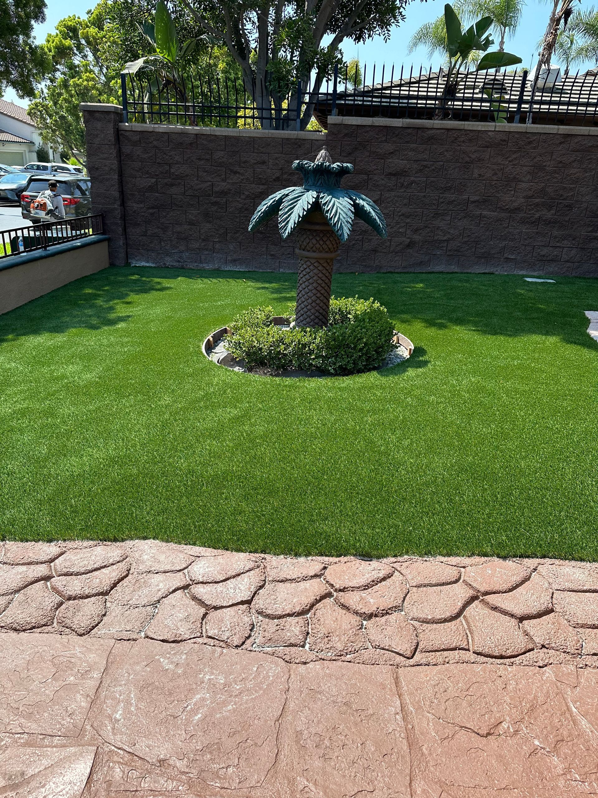 Green lawn with a decorative palm tree statue, bordered by a brick wall and paving.