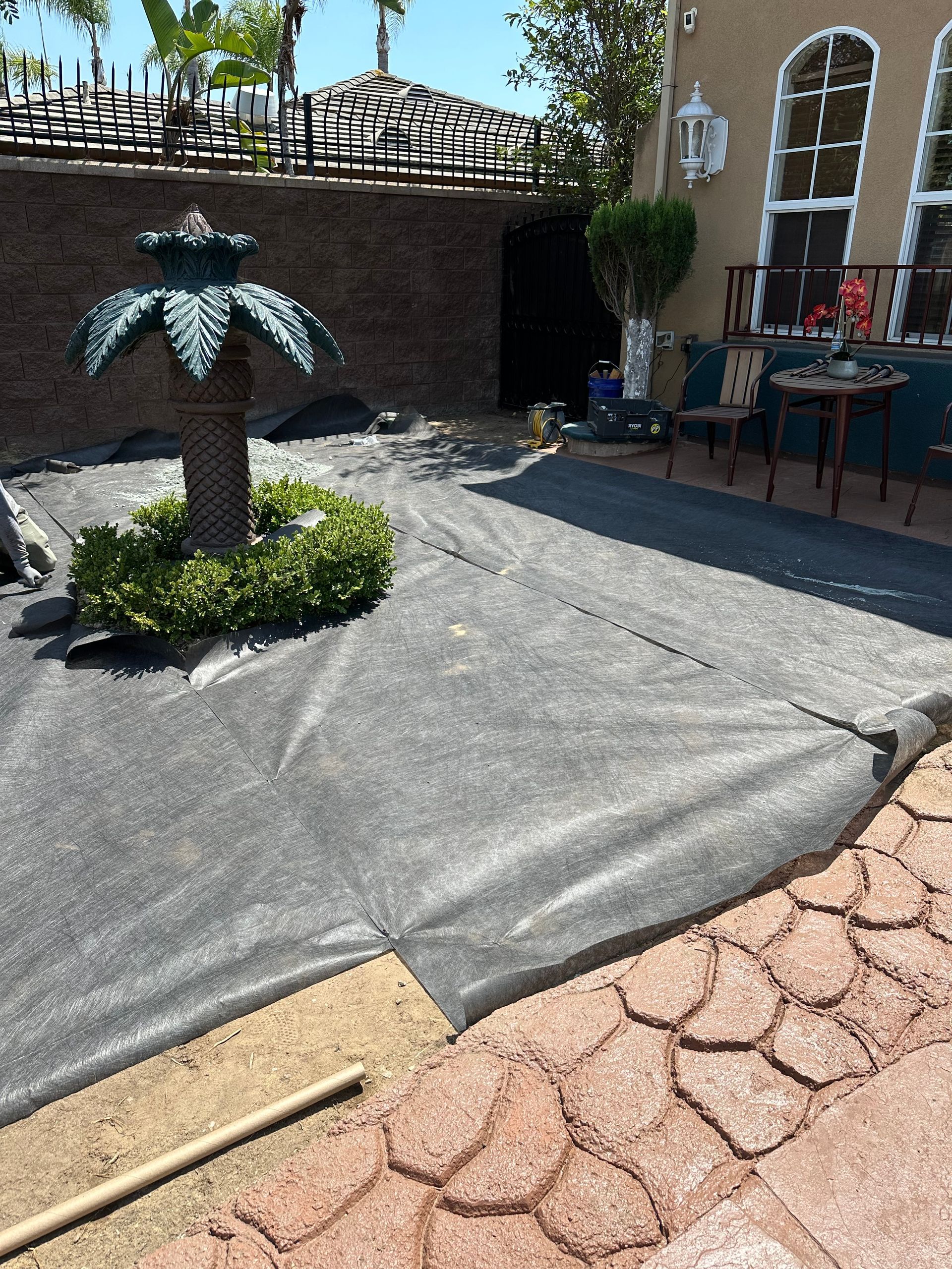 Black landscaping fabric covers a yard with a palm tree sculpture and brick patio.