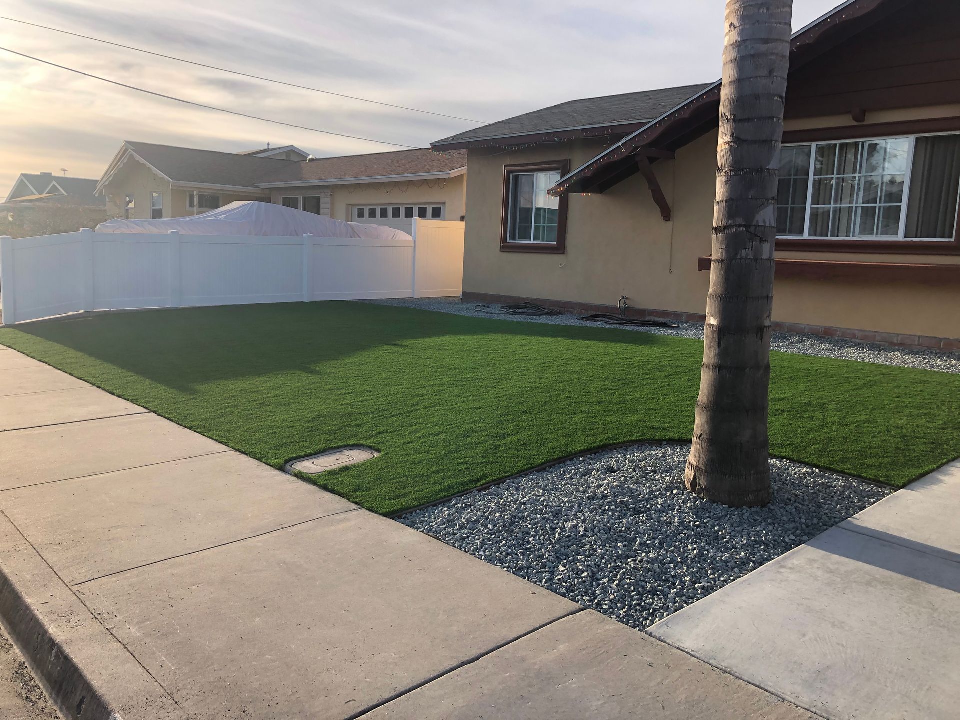 House with green lawn, white fence, and gray gravel. Sidewalk in foreground.