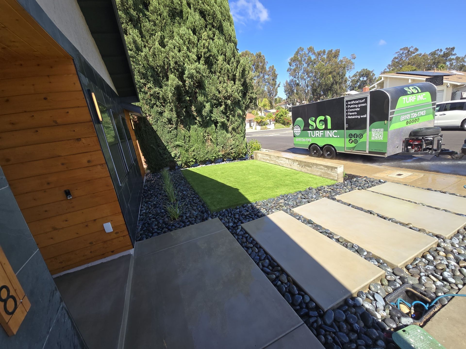 Modern home entrance with stone pavers, black pebbles, and green grass. A trailer is parked on the street.