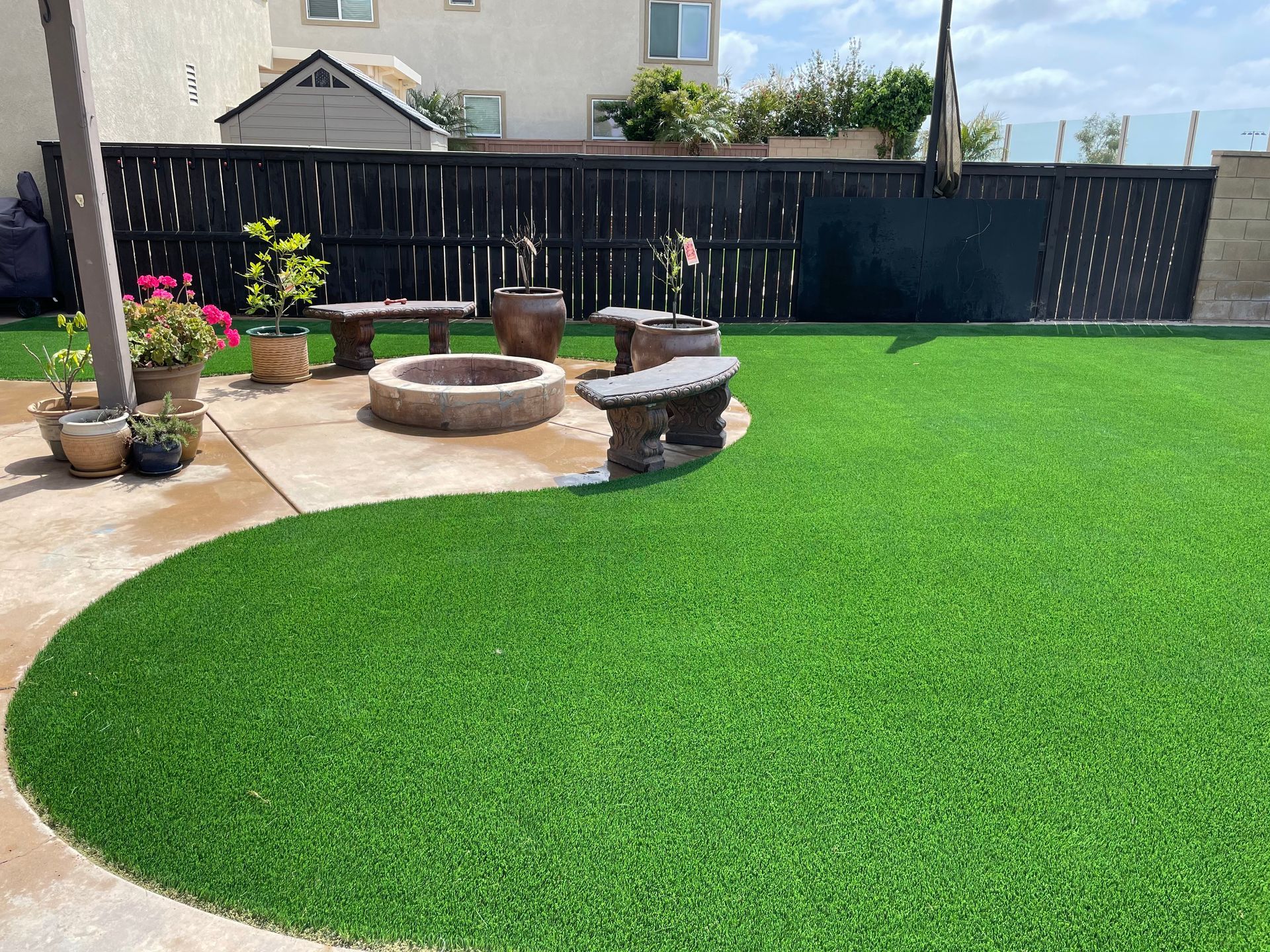 A backyard with green turf, a fire pit, stone benches, and potted plants next to a black fence.