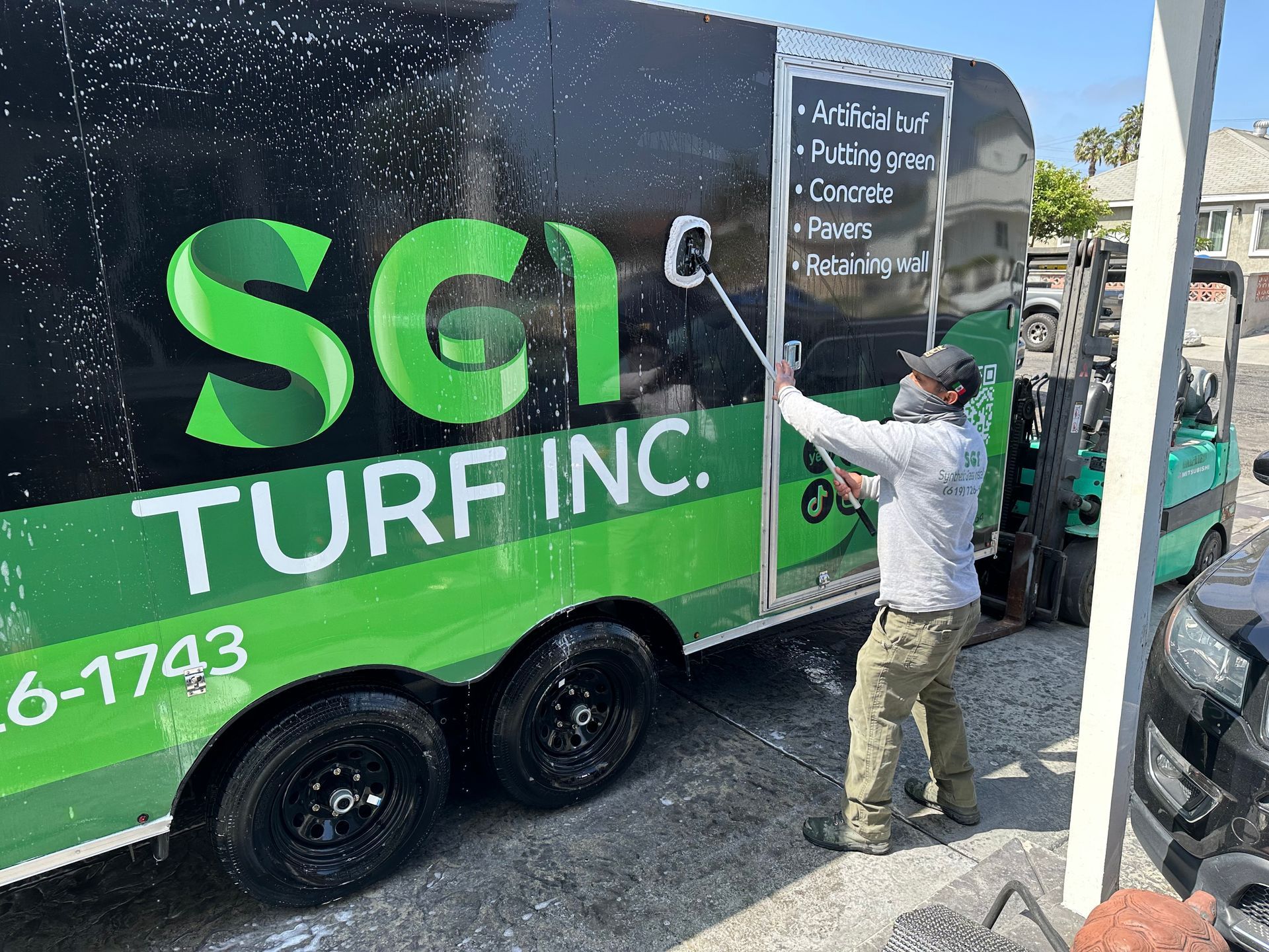 Man washing a green and black SCI Turf Inc. trailer with a foam brush. A forklift is visible nearby.