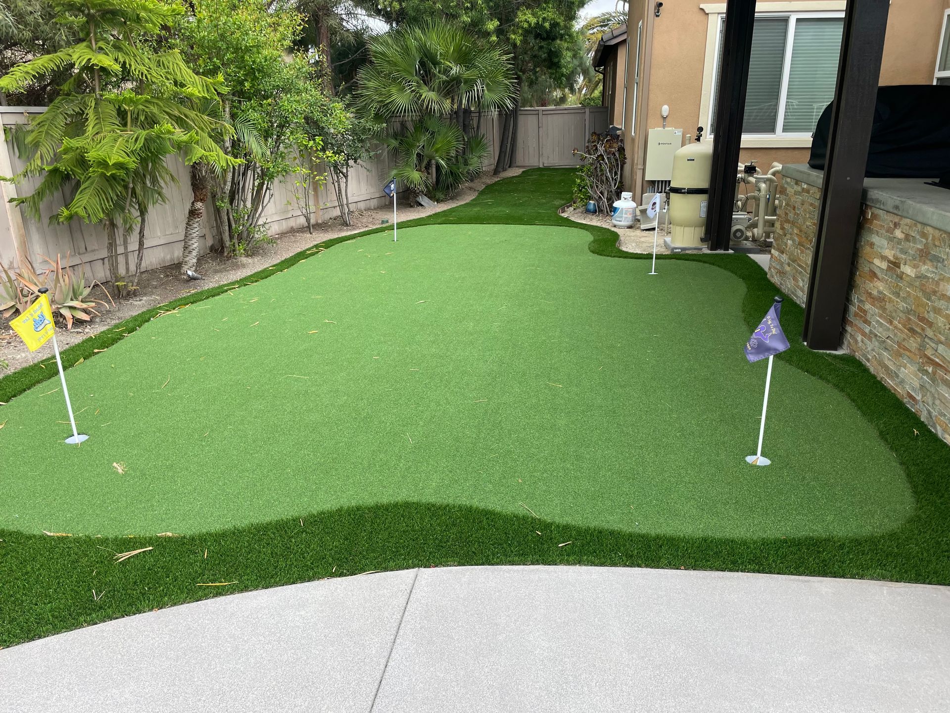 Backyard putting green with three flagsticks, surrounded by concrete and landscaping.