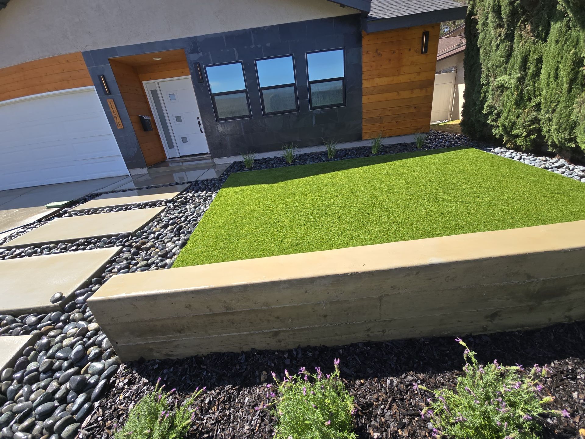 Modern home exterior with a green lawn bordered by black rocks and concrete pavers.