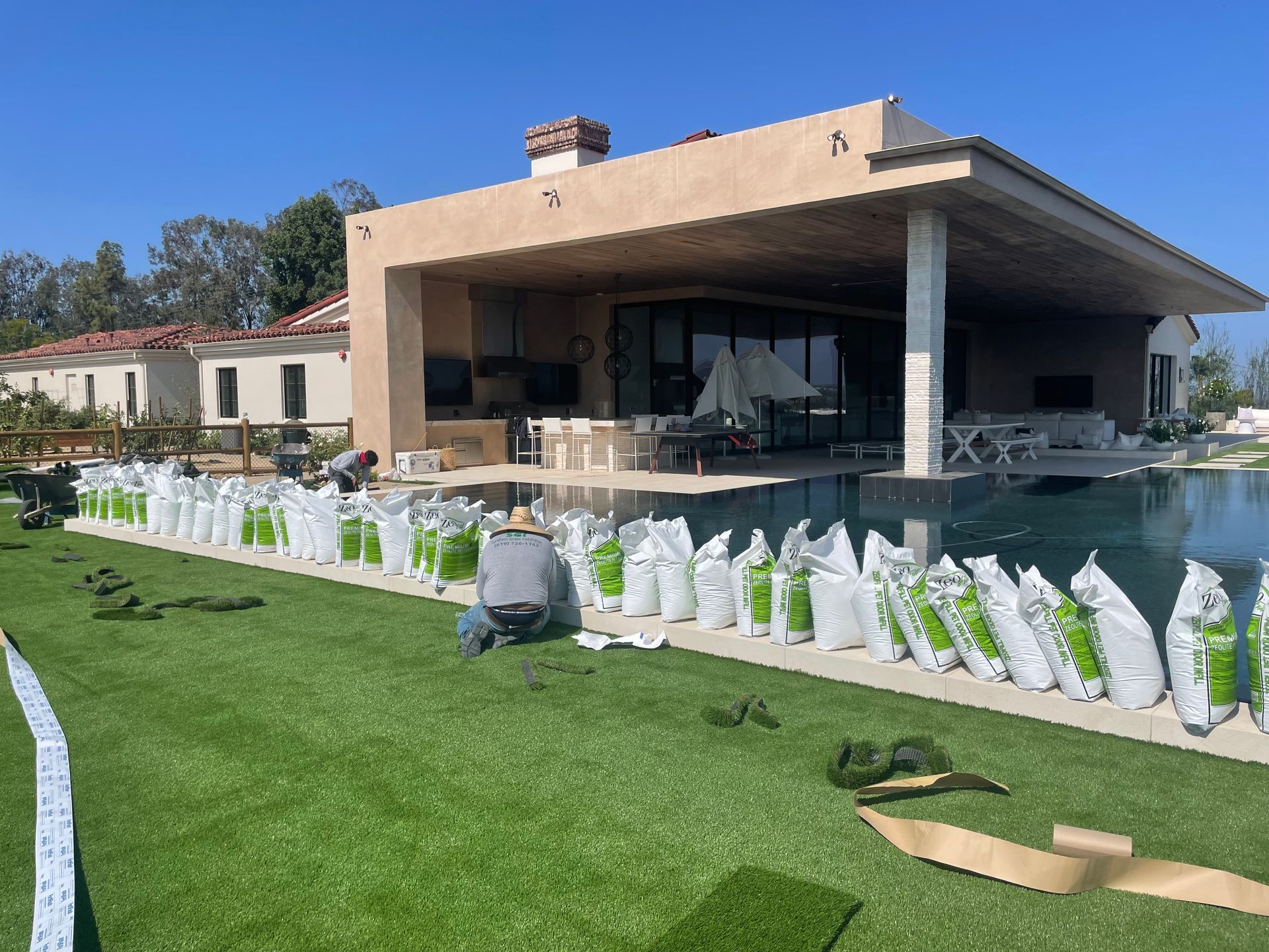 Workers installing artificial turf next to a pool at a luxury home. Green turf, beige house, blue sky.