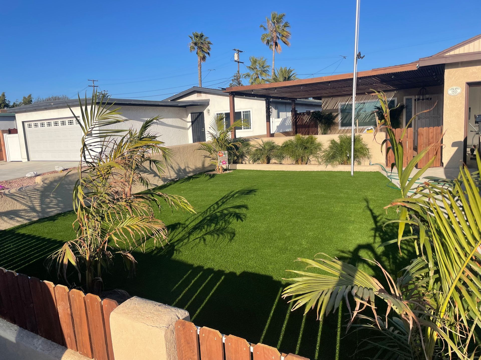 House exterior with artificial green lawn, plants, and wooden fence on a sunny day.