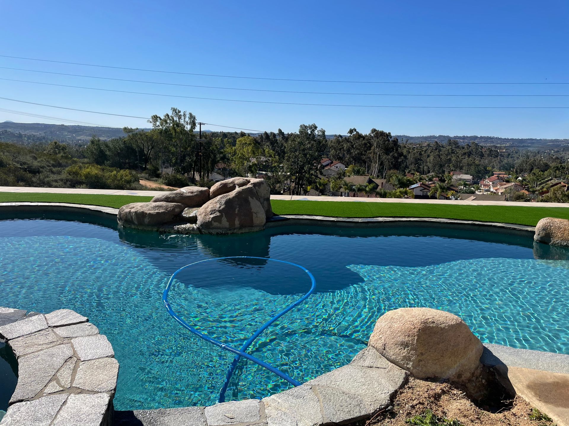 Swimming pool with clear blue water and rocky accents overlooks a scenic green landscape under a bright blue sky.