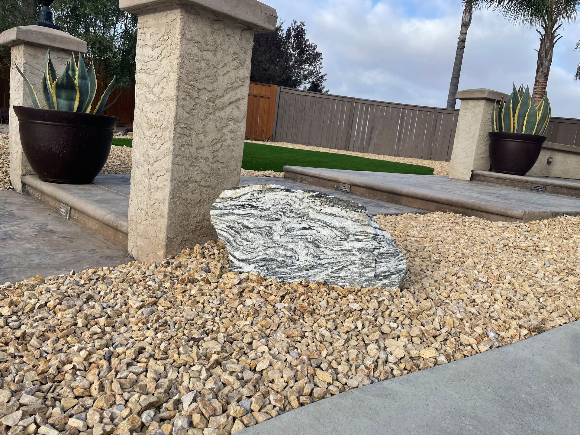 Large decorative rock on a bed of tan gravel, near a tan stucco pillar and potted plants.