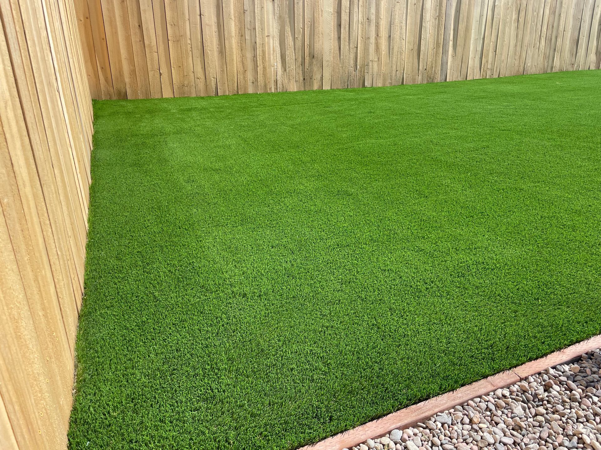 Green artificial turf in a fenced backyard, bordered by wood and gravel.
