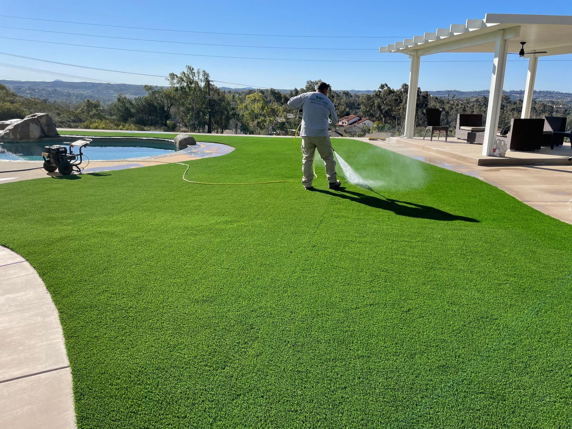 Man spraying water on green artificial turf lawn; pool and pergola in background.
