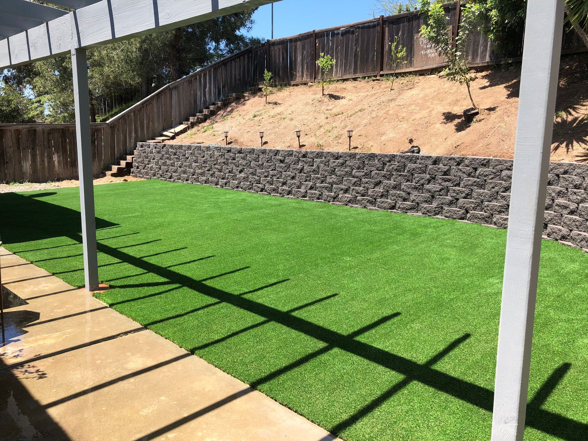 Backyard with artificial green turf, stone retaining wall, and wooden pergola casting a shadow.