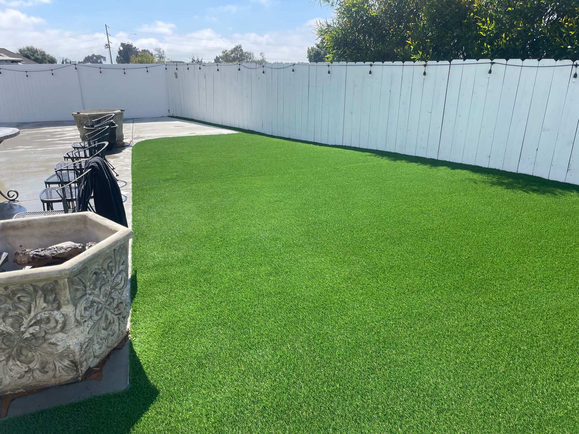 Green artificial turf in a backyard, bordered by a white fence and concrete patio. Sunny day.