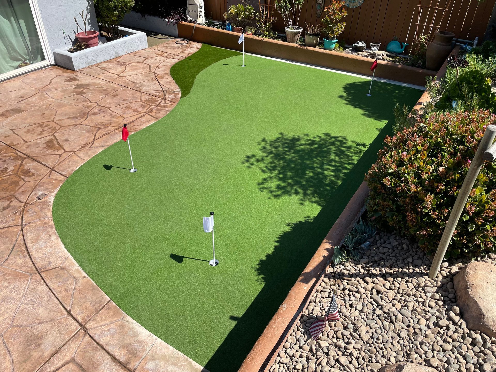 Artificial turf putting green in a backyard with multiple holes marked by flags, surrounded by landscaping.