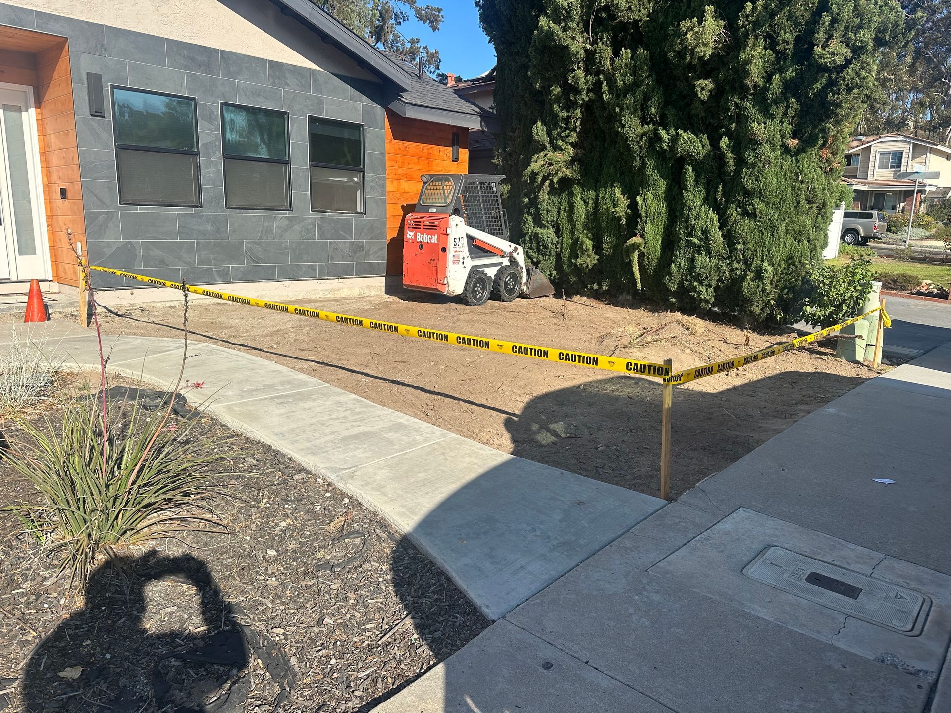 Bobcat in front yard excavation, caution tape surrounding work area. House with dark gray siding. Sunny day.
